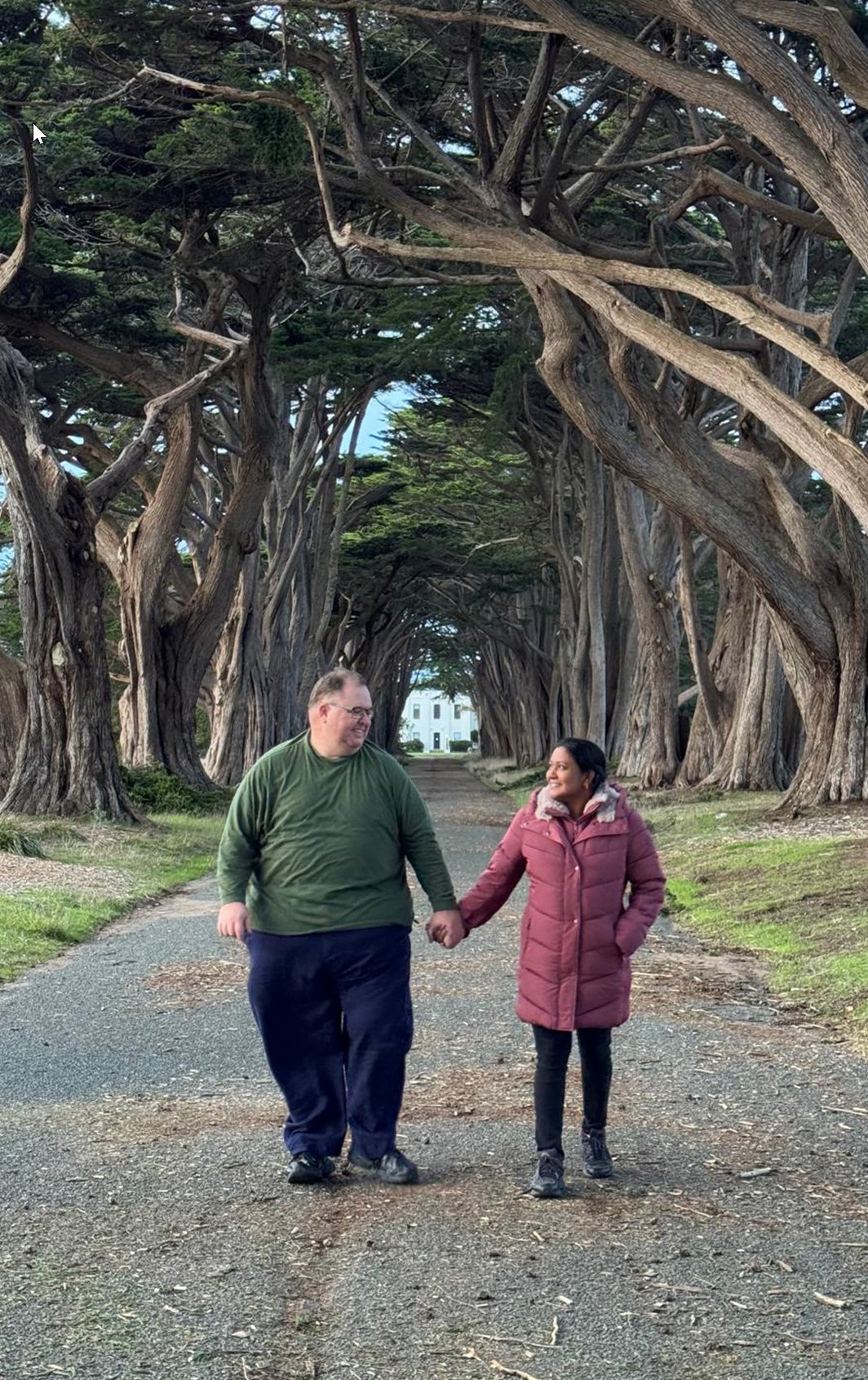 Andy and Shobha walking hand in hand under a canopy of cypress trees