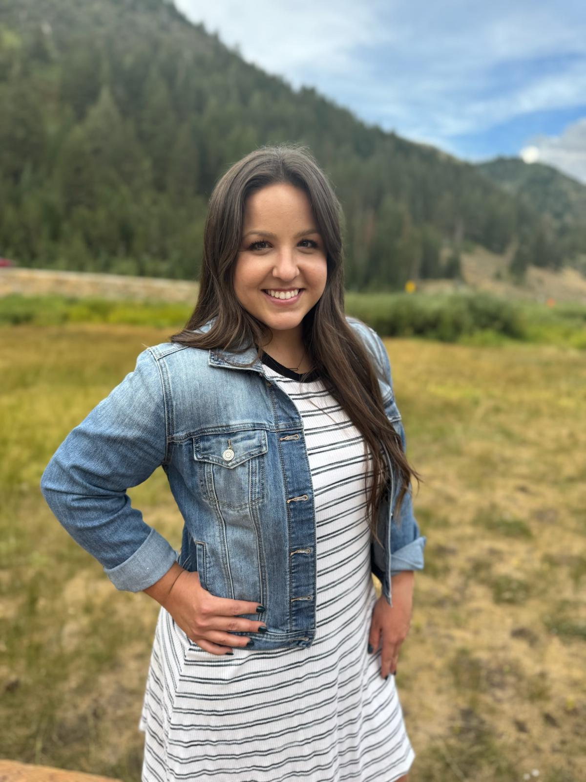 Young woman smiling in a denim jacket with green mountain meadow behind her