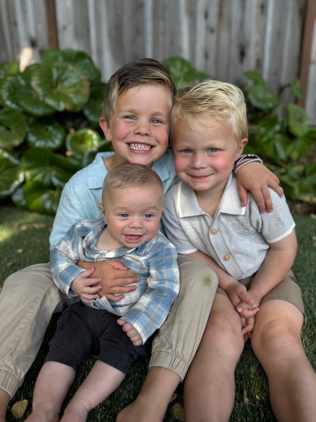 Three young brothers sitting together on the grass, the oldest holding the baby